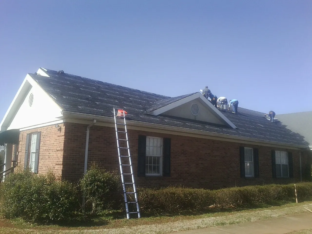 Crew tearing shingles off a brick ranch roof with a ladder set — Emergency Roof Repair work by our crew in Ledyard