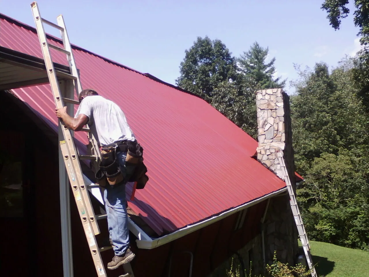 Roofer on a ladder beside a red metal roof and stone chimney — Soffit & Fascia Repair by Roofing Brothers in Ledyard